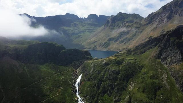 Scenic aerial view of Le Lac Bleu and a cascading waterfall surrounded by rugged mountains in the Pyrenees. Lush green valleys and dramatic peaks under a partly cloudy sky.