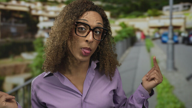 Trans woman raising middle finger at camera on street near sidewalk railing amid blurred city buildings; defiance.