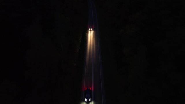 Aerial view of vehicles traveling along winding road at night, illuminated headlights cutting through darkness, showcasing dynamic motion and depth, camera follows action with smooth tracking shot