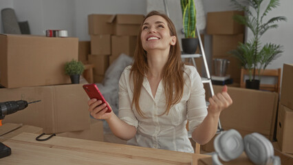 Fototapeta premium Woman holds smartphone during unpacking as moving boxes fill the room in new building; fresh start excitement.