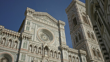 Fototapeta premium Cathedral rose window faces clear blue sky on building facade with adjacent bell tower, marble statues and ornate carvings; heritage serenity.