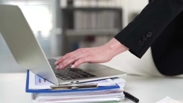 Unrecognizable businesswoman with manicured nails wearing a black suit working in the office, typing on a laptop keyboard resting on a stack of financial documents and charts