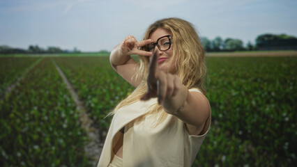 Woman with glasses and blonde hair gives thumbs up and points finger at camera in a rural forest field under blue sky; confidence.