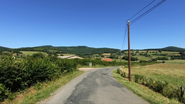 Scenic country road stretching through the rolling green hills and fields of the Morvan region in France under a clear blue sky.