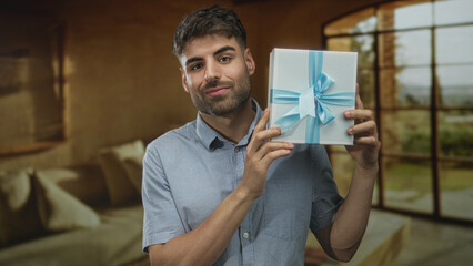 Fototapeta premium Young hispanic man holds blue ribboned gift box with hand and smiles in studio; celebration surprise.