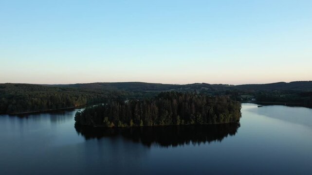 Aerial view of a wooded island surrounded by calm water at Lac des Settons, with lush green hills in the background under a clear evening sky.