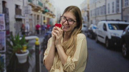 Obraz premium Young blonde woman wearing glasses and smiling while holding a small red heart on a busy street; love.