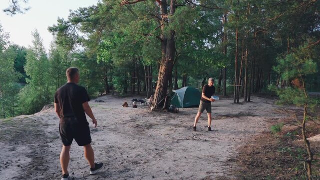Two individuals engage in casual disc throwing outdoors. Unwind in nature by tossing frisbee near camping gear. Relaxing evening of friendly disc tossing amidst wilderness and camping supplies