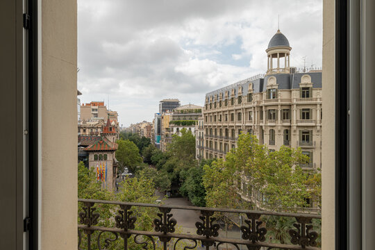 View from a balcony overlooking Barcelona Eixample district. Buildings with varied architecture and green trees are visible under a cloudy sky.