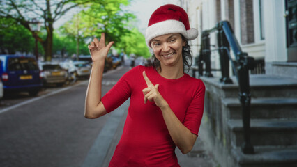 Fototapeta premium Woman in santa hat and red shirt points finger and gestures with both hands on a city street beside building steps while smiling broadly; holiday joy.