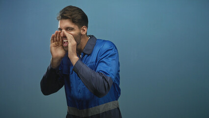 Hispanic man in blue work overalls cups hands to shout toward camera in studio; urgent call...
