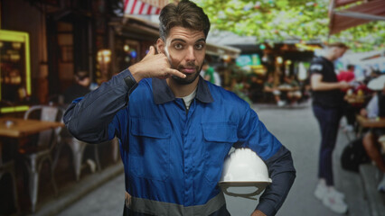 Obraz premium Hispanic man construction worker holding white hardhat makes call me hand gesture while wearing blue overalls in street; confidence readiness.