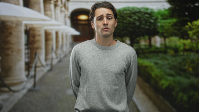 Man standing with hands behind back and exposed face, frowning gesture in building courtyard with columns and umbrella lined path; pensive solitude.