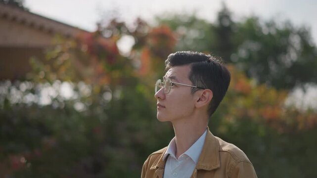 asian man outdoor portrait among shrubs, quiet student strolling and posing as street sketch subject, tan jacket and round glasses, multiple profile and back views, soft sunlight and bokeh, urban