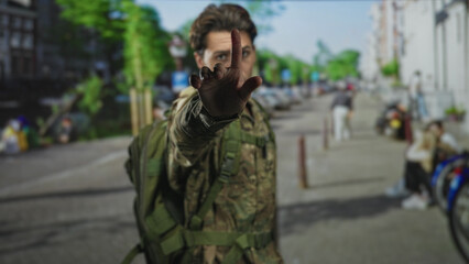 Obraz premium Man in camouflage uniform soldier with backpack raises hand, palm out toward camera on street lined with bicycles and buildings; duty tension.