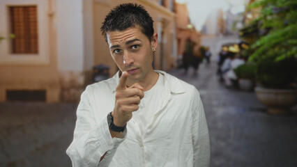 Naklejka premium Young man pointing in urban street setting provides a thoughtful gesture wearing white shirt amid blurred city backdrop capturing essence of contemplation in lively town environment.