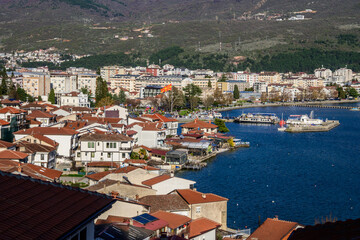 Obraz premium Panoramic cityscape of Ohrid, North Macedonia, showcasing red-tiled traditional architecture by the deep blue lake, bustling waterfront, and distant green mountains under bright daylight.