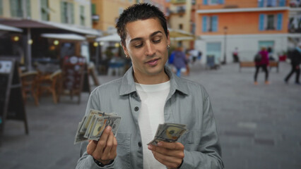 Naklejka premium Young hispanic man holding us dollars on a bustling restaurant terrace outdoors, exuding casual confidence amidst a lively coffee shop setting and vibrant street ambience.