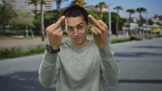 Young hispanic man in grey sweater shows both bare middlefinger gestures to camera on urban street; defiance.