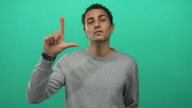 Hispanic man with defiant stare makes loser sign and points finger to camera in studio; mockery teasing.
