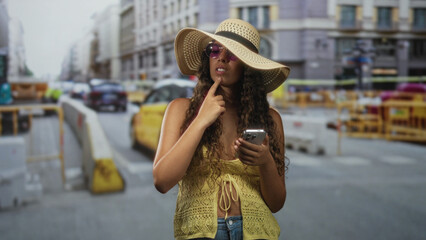 Hispanic woman holding smartphone and adjusting sun hat, hand raised in a hailing gesture on a busy city street with taxis and construction barriers  carefree summer travel. © Krakenimages.com