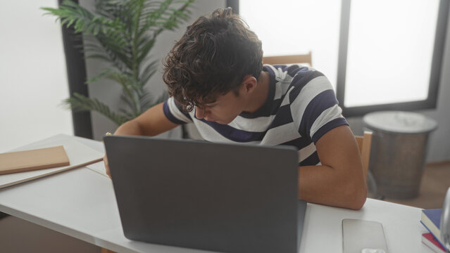 Teen student leans over laptop writing with pencil at studio desk, head bent and arm resting on table; study concentration.