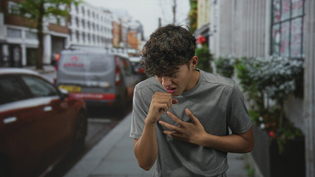Teenage boy in grey t shirt clutching throat and coughing on a busy city street beside parked cars and a delivery van; distress urgency.