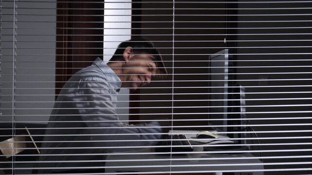 Young man experiencing stress and burnout, angrily hitting his computer screen and keyboard, seen through venetian blinds in a dimly lit office environment