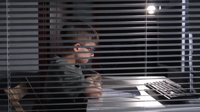 Young boy wearing glasses intently reading a book at a desk while a computer keyboard sits nearby, viewed through venetian blinds in a dimly lit room representing focus and education