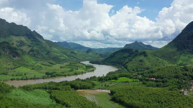 Scenic view of a winding river surrounded by green mountains and fields in the countryside between Luang Prabang and Nong Khiaw, Laos. Dramatic clouds fill the sky above the vibrant landscape.