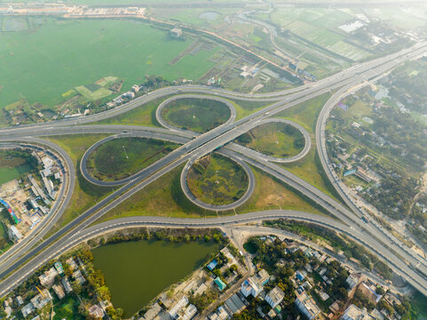 Aerial view of a cloverleaf interchange weaving amongst urban structures and a tranquil pond, contrasting the green fields, Bhanga, Bangladesh.