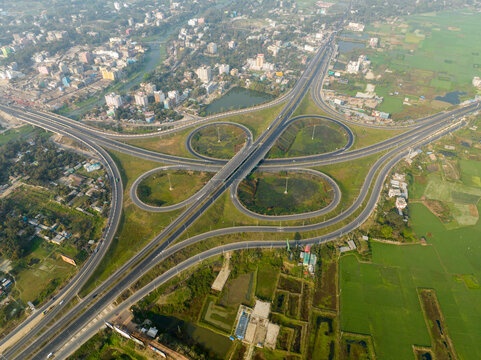 Aerial view of the Bhanga Interchange, a cloverleaf design of roads weaving amidst a tapestry of green fields and urban development, Bhanga, Dhaka Division, Bangladesh.