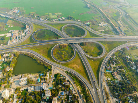 Aerial view of a cloverleaf interchange with smooth curves of asphalt cutting through a landscape of verdant fields and dense urban development, Bhanga, Dhaka Division, Bangladesh.