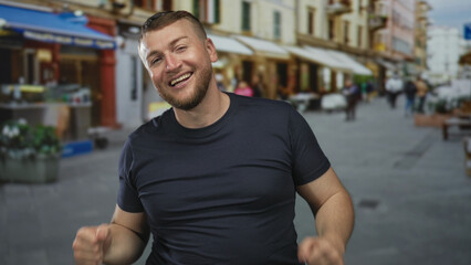 Fototapeta premium Man young caucasian smiling with fists raised, bare forearms visible in a busy street market wearing a dark t shirt; joy celebration.
