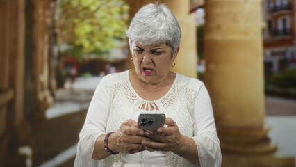 Fototapeta premium Woman holds smartphone, frowns at the screen near a colonnade outside a building on a street; frustration technology problem.