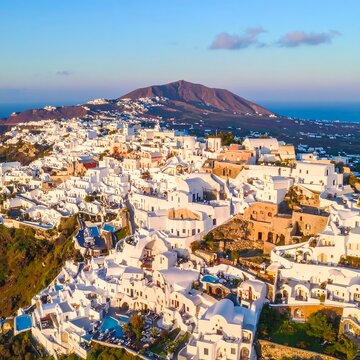 Aerial view of a picturesque coastal village with whitewashed buildings, nestled against a backdrop of a volcano and the serene sea at dusk