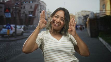 Fototapeta premium Hispanic woman smiling with hands raised in playful pose wearing striped v neck shirt on a cobblestone street near buildings; joyful playfulness.