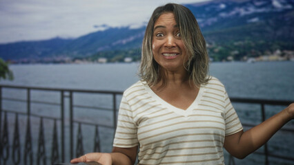 Fototapeta premium Woman holding smartphone and smiling while shrugging at railing on street by lake and mountains; delight curiosity.