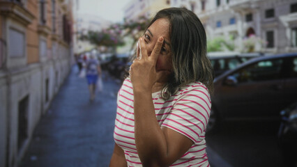 Woman covers face with hand in striped shirt on street, hand over eyes beside parked car and buildings; embarrassment.