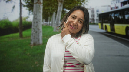 Fototapeta premium Hispanic middle aged woman with closed eyes touching her cheek with fingers beside a yellow city bus on street near tree lined sidewalk; contemplation.