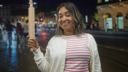 Woman smiling holds rolling pin upright with right hand wearing striped shirt and cardigan on a busy city street at night  joyful confidence. © Krakenimages.com