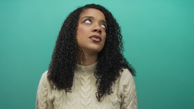 African american woman rolling eyes against teal background in studio wearing cream cable knit sweater; boredom.