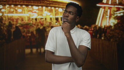 Obraz premium Man wearing glasses touches chin in pensive pose at street fair under glowing night lights; contemplation.
