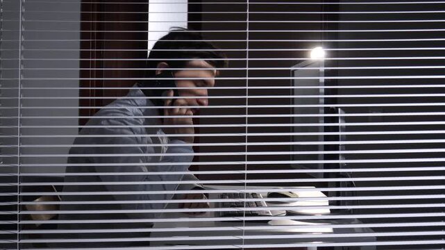 Man talking on a phone, working at a computer in a dark office, viewed through window blinds, highlighting concepts of privacy, espionage, secret communication, and surveillance