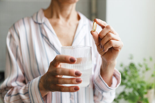 Hands of middle-aged woman holding transparent glass of water and pill of polyunsaturated fatty acids or vitamin D3. Unrecognizable mature female taking dietary supplements in morning to stay healthy