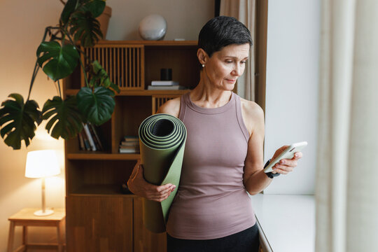 Woman of 50s in perfect shape standing in living-room in sports clothes holding rolled mat and smartphone, getting ready for group training online via video application on her phone