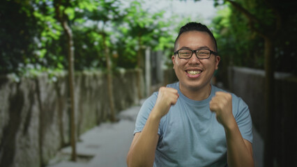 Man with fists raised and mouth open shouting on a street lined with trees and stone walls, wearing glasses and a light blue t shirt  joy celebration. © Krakenimages.com