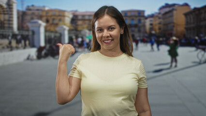 Fototapeta premium Woman points thumb over shoulder on a busy city street showing thumb gesture and smile, casual t shirt visible; confidence travel.
