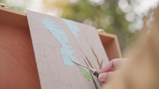 caucasian woman applying thick paint to canvas with palette knife, creating impasto sky and branch texture on portable easel in sunlit garden, closeup hand movements with warm bokeh background,