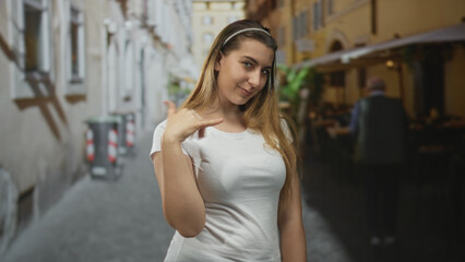 Woman pointing thumb to herself and making a phone gesture while smiling in a narrow street cafe; playful confidence.